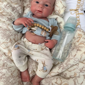 May include: A realistic baby doll wearing a light blue onesie and cream-colored pants with a Peter Rabbit print. The doll has a pacifier and a baby bottle. The doll is lying on a patterned blanket.