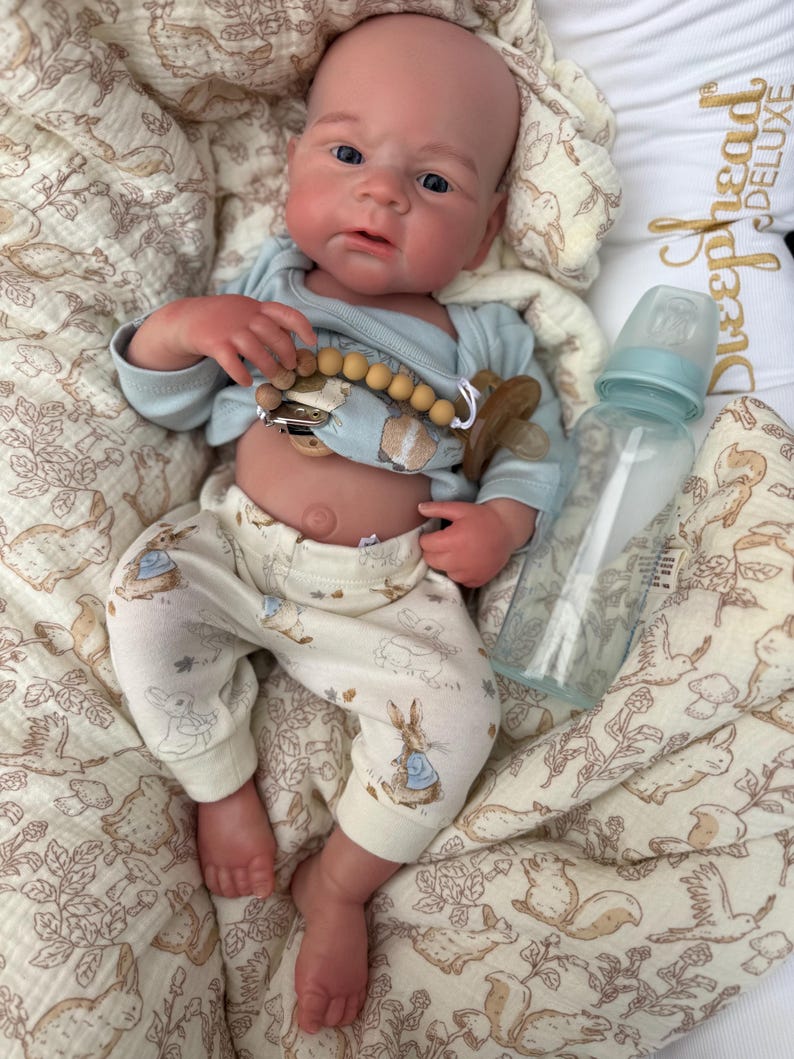 May include: A realistic baby doll wearing a light blue onesie and cream-colored pants with a Peter Rabbit print. The doll has a pacifier and a baby bottle. The doll is lying on a patterned blanket.