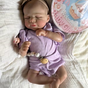 Pu&ograve; includere: Una bambola realistica di neonato con gli occhi chiusi, che indossa una tutina a maniche corte color viola chiaro e una fascia color marrone chiaro. Una clip per ciuccio bianca con perline di legno &egrave; attaccata alla tutina. Accanto alla bambola c'&egrave; un cartello rotondo con la scritta Welcome Baby Girl.