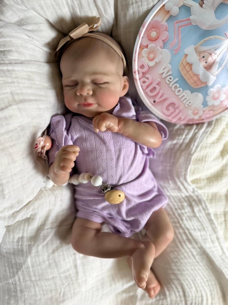 Pu&ograve; includere: Una bambola realistica di neonato con gli occhi chiusi, che indossa una tutina a maniche corte color viola chiaro e una fascia color marrone chiaro. Una clip per ciuccio bianca con perline di legno &egrave; attaccata alla tutina. Accanto alla bambola c'&egrave; un cartello rotondo con la scritta Welcome Baby Girl.