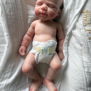 May include: A realistic newborn baby doll with light skin, closed eyes, and a serene expression. The doll has detailed facial features, soft limbs, and is wearing a white disposable diaper with yellow adhesive tabs. The doll is lying on its back.