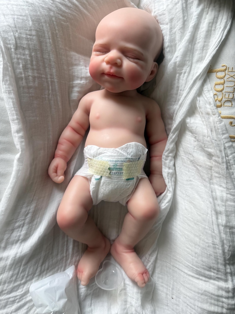 May include: A realistic newborn baby doll with light skin, closed eyes, and a serene expression. The doll has detailed facial features, soft limbs, and is wearing a white disposable diaper with yellow adhesive tabs. The doll is lying on its back.