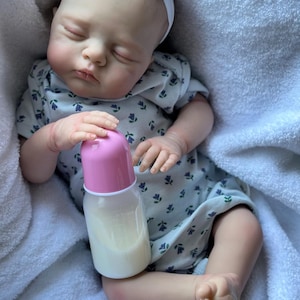 May include: A realistic reborn doll with closed eyes, wearing a white short-sleeved onesie with a small blue floral pattern and a white headband with a flower. The doll is holding a baby bottle with a pink cap and a white liquid inside.