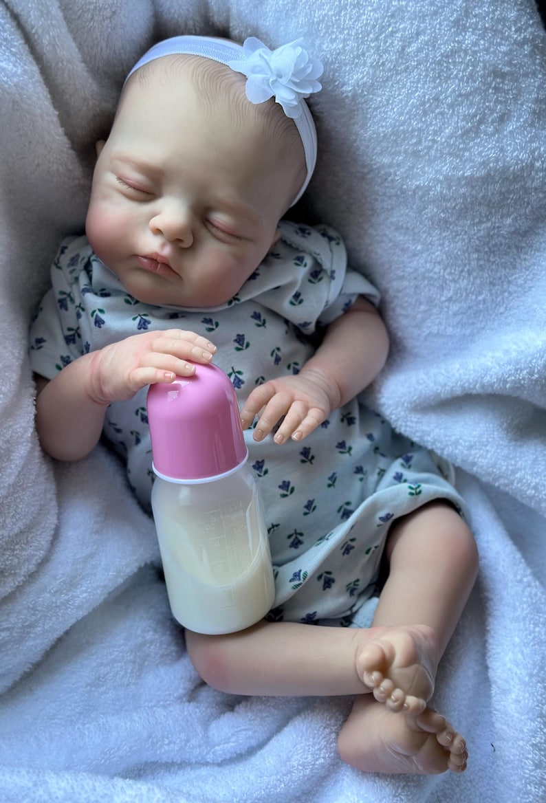May include: A realistic reborn doll with closed eyes, wearing a white short-sleeved onesie with a small blue floral pattern and a white headband with a flower. The doll is holding a baby bottle with a pink cap and a white liquid inside.