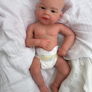 K&ouml;nnte beinhalten: Eine realistische Neugeborenen-Puppe mit heller Haut, einer Glatze und dunklen Augen. Die Puppe hat detaillierte Gesichtsz&uuml;ge, einschlie&szlig;lich feiner Augenbrauen und eines sanften Ausdrucks, und tr&auml;gt eine wei&szlig;e Einwegwindel mit gelben Klebestreifen.