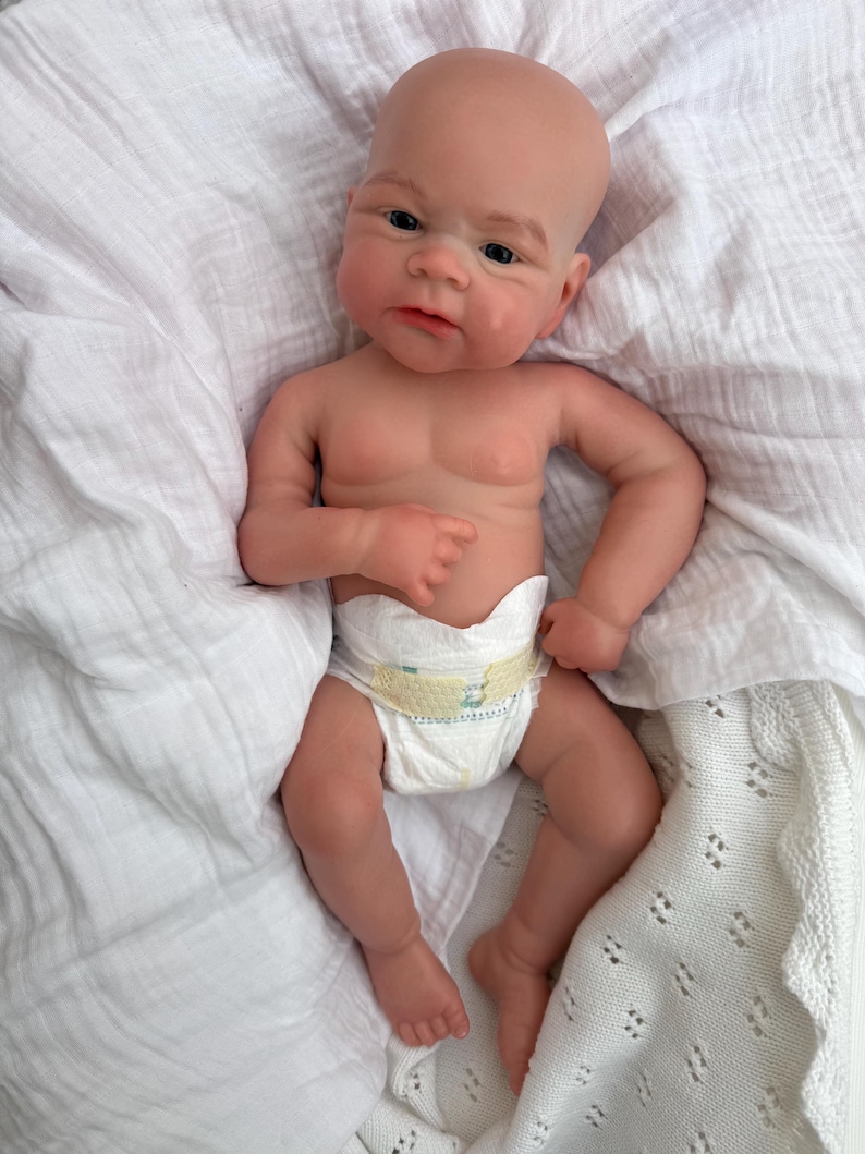 K&ouml;nnte beinhalten: Eine realistische Neugeborenen-Puppe mit heller Haut, einer Glatze und dunklen Augen. Die Puppe hat detaillierte Gesichtsz&uuml;ge, einschlie&szlig;lich feiner Augenbrauen und eines sanften Ausdrucks, und tr&auml;gt eine wei&szlig;e Einwegwindel mit gelben Klebestreifen.
