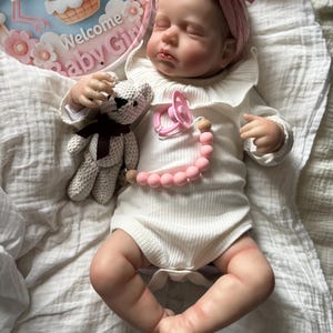 May include: A realistic baby doll with closed eyes, wearing a white ribbed long-sleeve bodysuit with a ruffled collar and a pink knotted headband. The doll holds a small crocheted bear and has a pink pacifier attached to a pink and wood bead chain.