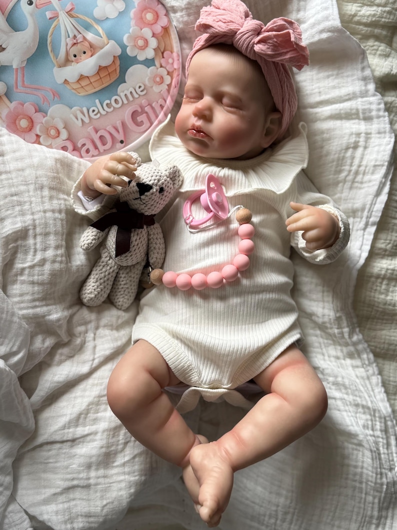 May include: A realistic baby doll with closed eyes, wearing a white ribbed long-sleeve bodysuit with a ruffled collar and a pink knotted headband. The doll holds a small crocheted bear and has a pink pacifier attached to a pink and wood bead chain.