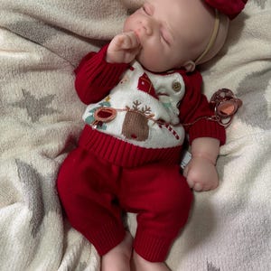 Pode incluir: Uma boneca beb&eacute; a dormir com tom de pele claro, vestindo uma camisola de malha vermelha e branca com um padr&atilde;o de rena e &aacute;rvore de Natal, cal&ccedil;as de malha vermelhas a combinar e uma fita com um la&ccedil;o vermelho grande. Uma chupeta em ouro rosa est&aacute; presa &agrave; camisola.