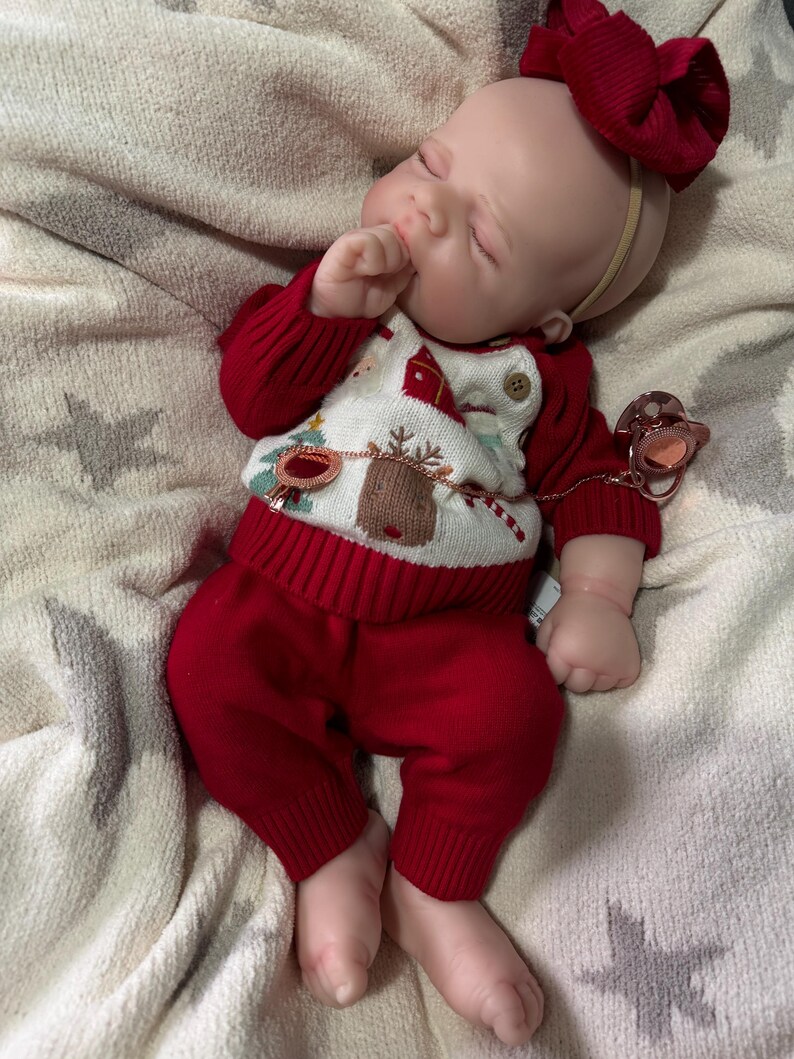 Pode incluir: Uma boneca beb&eacute; a dormir com tom de pele claro, vestindo uma camisola de malha vermelha e branca com um padr&atilde;o de rena e &aacute;rvore de Natal, cal&ccedil;as de malha vermelhas a combinar e uma fita com um la&ccedil;o vermelho grande. Uma chupeta em ouro rosa est&aacute; presa &agrave; camisola.