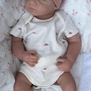 Może przedstawiać: Realistyczna lalka dla dziecka z zamkniętymi oczami, w r&oacute;żowej opasce na głowę i białym body ozdobionym małymi r&oacute;żowymi chmurkami. Lalka leży na kocu we wzory kwiatowe, tworząc miękką i kojącą scenę.