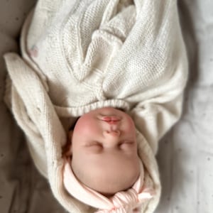 May include: A realistic newborn doll with a pale complexion and rosy cheeks, wrapped in a cream-colored cable knit blanket. The doll features closed eyes, a small nose, and pink lips, and is wearing a soft, light pink knotted headband on its head.