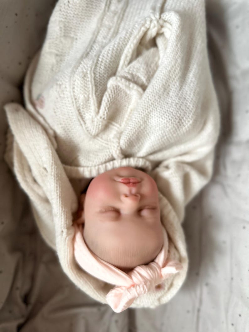 May include: A realistic newborn doll with a pale complexion and rosy cheeks, wrapped in a cream-colored cable knit blanket. The doll features closed eyes, a small nose, and pink lips, and is wearing a soft, light pink knotted headband on its head.