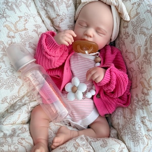 May include: A baby doll wearing a pink cardigan, striped onesie, and a cream-colored bow headband. A clear baby bottle and a pacifier with a flower clip are also visible. The doll is lying on a patterned blanket.
