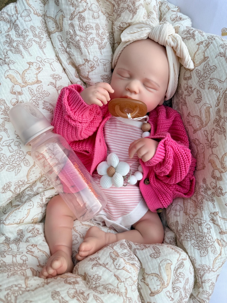 May include: A baby doll wearing a pink cardigan, striped onesie, and a cream-colored bow headband. A clear baby bottle and a pacifier with a flower clip are also visible. The doll is lying on a patterned blanket.