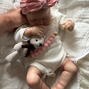 May include: A realistic baby doll with light skin wearing a white ribbed long-sleeve onesie and a dusty pink fabric bow headband. The doll holds a small crocheted beige teddy bear with a brown ribbon and a pink silicone bead pacifier clip.