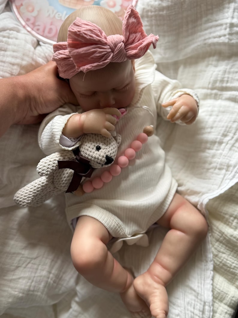 May include: A realistic baby doll with light skin wearing a white ribbed long-sleeve onesie and a dusty pink fabric bow headband. The doll holds a small crocheted beige teddy bear with a brown ribbon and a pink silicone bead pacifier clip.