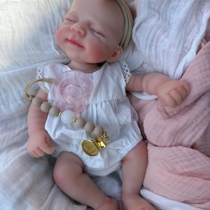 May include: A realistic baby doll with closed eyes and a soft expression, wearing a white sleeveless romper with lace trim. The doll has a beige bow headband, a pink pacifier, and a pacifier clip featuring beige and white beads with a gold metal clasp.