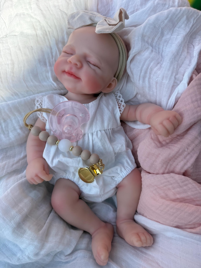May include: A realistic baby doll with closed eyes and a soft expression, wearing a white sleeveless romper with lace trim. The doll has a beige bow headband, a pink pacifier, and a pacifier clip featuring beige and white beads with a gold metal clasp.