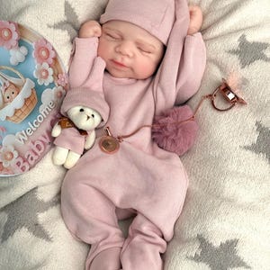 Pu&ograve; includere: Una bambola realistica che indossa una tutina a maniche lunghe a coste color rosa polvere e un berretto abbinato. La bambola tiene in mano un piccolo orsetto di peluche bianco con berretto e sciarpa rosa. &Egrave; attaccato un ciuccio oro rosa con catenella.