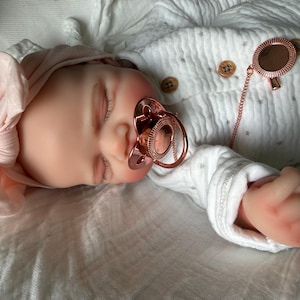 May include: A realistic baby doll with closed eyes, wearing a light pink knotted headband and a white long-sleeved onesie with a subtle grey star pattern and brown buttons. The doll has a rose gold pacifier and a matching rose gold pacifier clip.