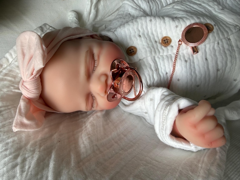 May include: A realistic baby doll with closed eyes, wearing a light pink knotted headband and a white long-sleeved onesie with a subtle grey star pattern and brown buttons. The doll has a rose gold pacifier and a matching rose gold pacifier clip.