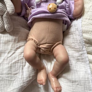 Pu&ograve; includere: Una bambola realistica indossa un top a maniche corte color viola chiaro con un motivo testurizzato e un pannolino beige. Una catenella portaciuccio con perline bianche e beige e un ciuccio rotondo beige &egrave; attaccata al top. Un giocattolo all'uncinetto grigio &egrave; visibile.