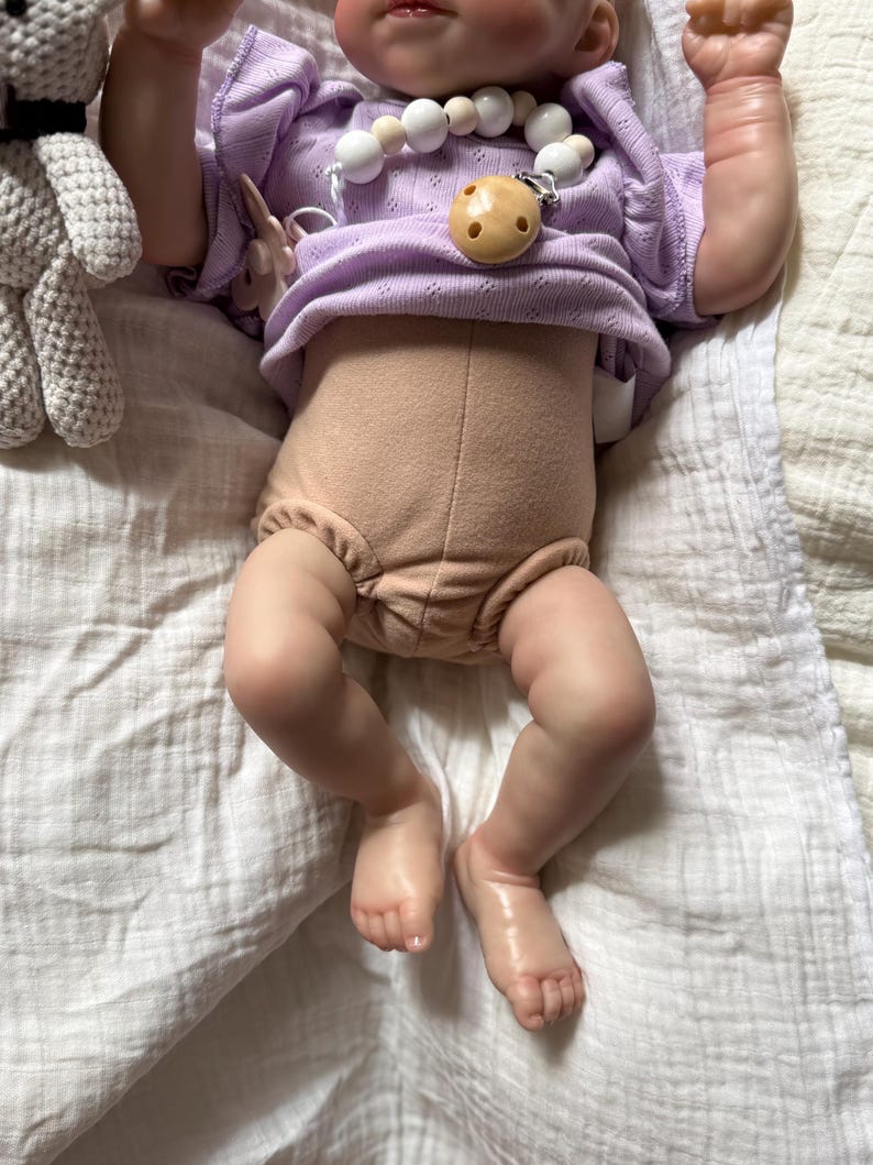 Pu&ograve; includere: Una bambola realistica indossa un top a maniche corte color viola chiaro con un motivo testurizzato e un pannolino beige. Una catenella portaciuccio con perline bianche e beige e un ciuccio rotondo beige &egrave; attaccata al top. Un giocattolo all'uncinetto grigio &egrave; visibile.