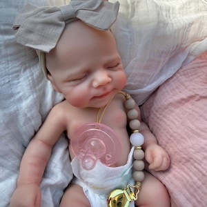 May include: A realistic newborn doll with closed eyes and a light skin tone, wearing a beige fabric bow headband and a white diaper. The doll has a pink pacifier attached to a beaded pacifier clip featuring matte taupe beads and a gold metal clasp.