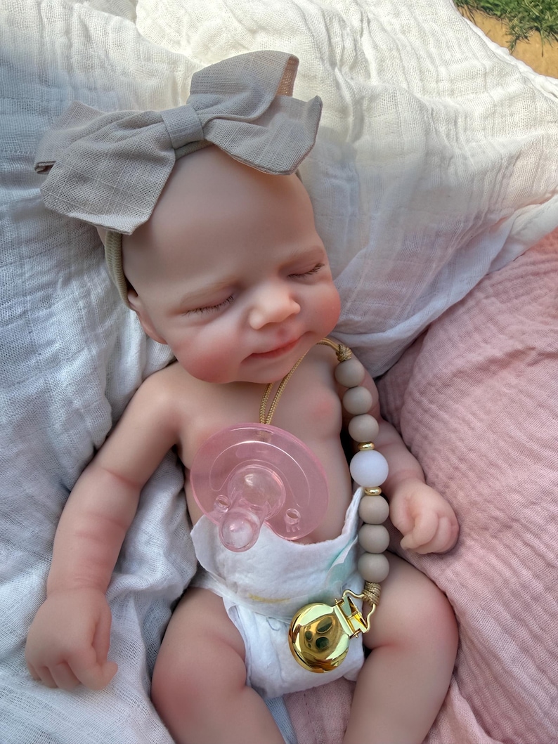 May include: A realistic newborn doll with closed eyes and a light skin tone, wearing a beige fabric bow headband and a white diaper. The doll has a pink pacifier attached to a beaded pacifier clip featuring matte taupe beads and a gold metal clasp.