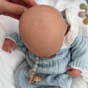 K&ouml;nnte beinhalten: Eine Babypuppe mit einem glatten, hellen Kopf, bekleidet mit einem hellblauen Zopfmuster-Pullover mit wei&szlig;em Kragen. Die Puppe hat einen Schnuller an einer Kette mit grauen und h&ouml;lzernen Perlen. Die Puppe liegt auf einem wei&szlig;en Stoff.