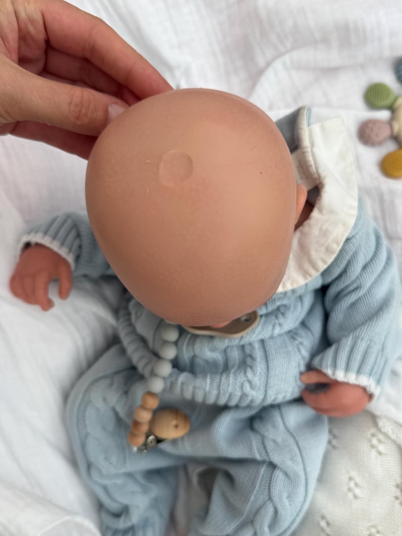 K&ouml;nnte beinhalten: Eine Babypuppe mit einem glatten, hellen Kopf, bekleidet mit einem hellblauen Zopfmuster-Pullover mit wei&szlig;em Kragen. Die Puppe hat einen Schnuller an einer Kette mit grauen und h&ouml;lzernen Perlen. Die Puppe liegt auf einem wei&szlig;en Stoff.