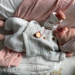 May include: A realistic baby doll with light skin, wearing a white textured long-sleeve shirt and matching pants with small brown buttons. The doll has a pink headband, a rose gold pacifier in its mouth, and a small rose gold circular pendant necklace.