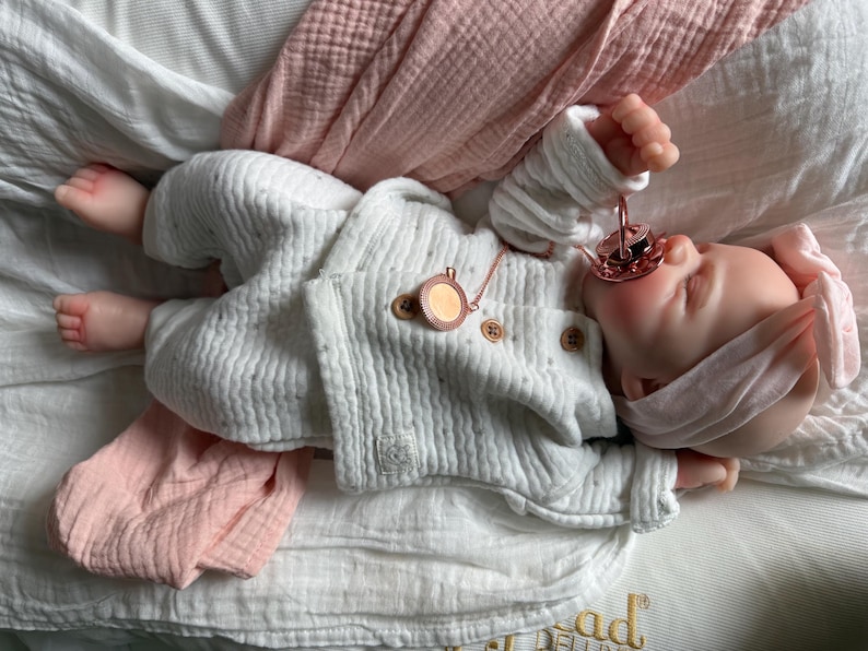 May include: A realistic baby doll with light skin, wearing a white textured long-sleeve shirt and matching pants with small brown buttons. The doll has a pink headband, a rose gold pacifier in its mouth, and a small rose gold circular pendant necklace.