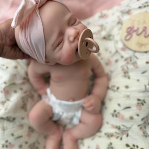 May include: A realistic newborn baby doll with closed eyes, wearing a light pink knotted headband and a beige pacifier. The doll has a fair complexion and is dressed in a white nappy. A wooden disc with the text It's a girl is visible nearby.