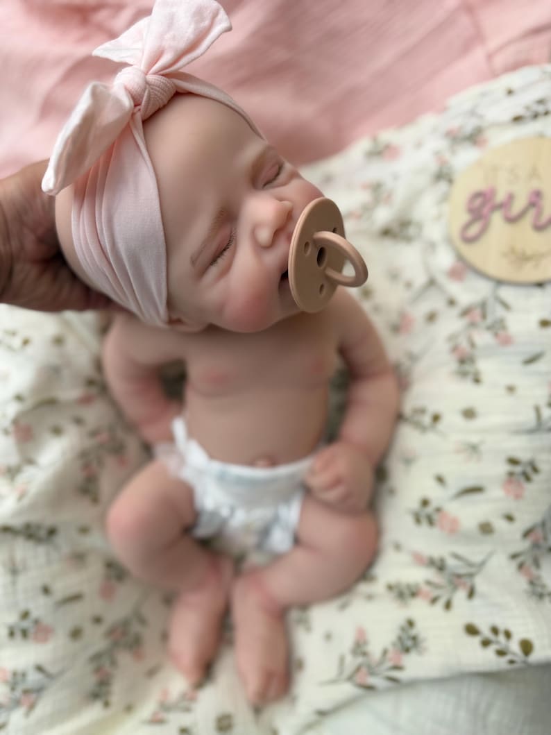 May include: A realistic newborn baby doll with closed eyes, wearing a light pink knotted headband and a beige pacifier. The doll has a fair complexion and is dressed in a white nappy. A wooden disc with the text It's a girl is visible nearby.