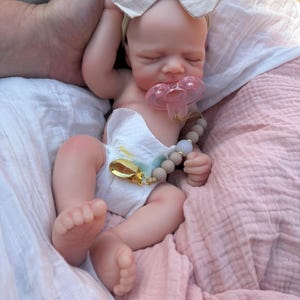 May include: A realistic silicone baby doll with closed eyes, wearing a white diaper and a beige fabric bow headband. The doll has a pink pacifier in its mouth attached to a beige beaded pacifier clip with a gold-toned metal clasp.