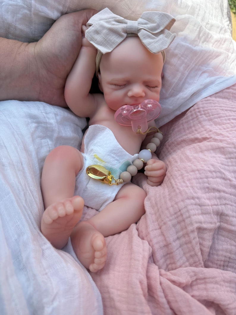May include: A realistic silicone baby doll with closed eyes, wearing a white diaper and a beige fabric bow headband. The doll has a pink pacifier in its mouth attached to a beige beaded pacifier clip with a gold-toned metal clasp.