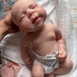 Pu&ograve; includere: Una bambola realistica di neonato con pelle pallida, occhi chiusi e un'espressione dolce. La bambola ha una testa liscia e senza capelli e indossa un pannolino usa e getta bianco. La bambola &egrave; posizionata in una postura reclinata con braccia e gambe piegate.