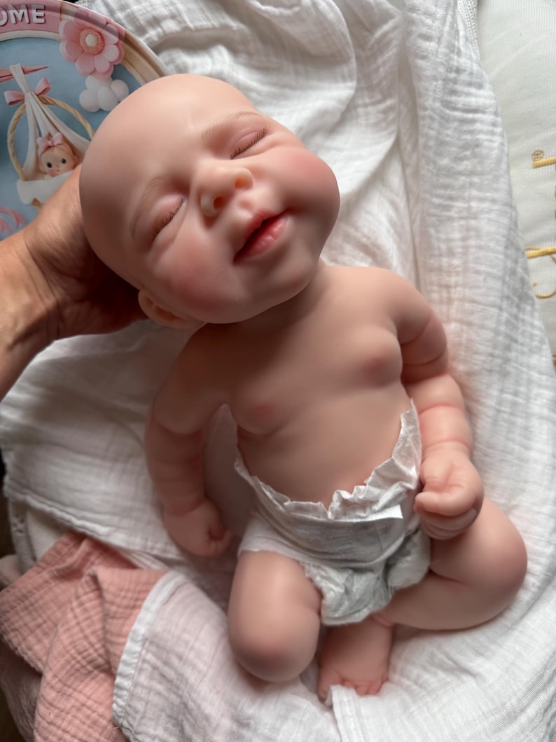 Pu&ograve; includere: Una bambola realistica di neonato con pelle pallida, occhi chiusi e un'espressione dolce. La bambola ha una testa liscia e senza capelli e indossa un pannolino usa e getta bianco. La bambola &egrave; posizionata in una postura reclinata con braccia e gambe piegate.