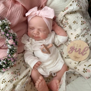 May include: A realistic newborn doll with closed eyes, wearing a white ribbed long-sleeve onesie and a light pink bow headband. The doll has a tan pacifier on its chest and is wrapped in a floral muslin blanket. A wooden disc reads IT'S A girl.