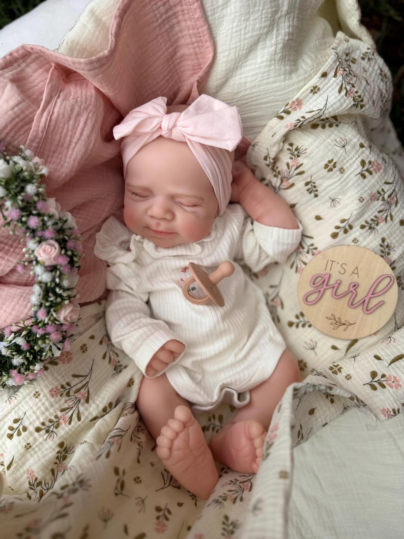 May include: A realistic newborn doll with closed eyes, wearing a white ribbed long-sleeve onesie and a light pink bow headband. The doll has a tan pacifier on its chest and is wrapped in a floral muslin blanket. A wooden disc reads IT'S A girl.