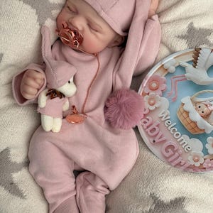 Pu&ograve; includere: Una bambola realistica che indossa una tutina rosa a coste a maniche lunghe con piedini e un berretto abbinato. La bambola ha un ciuccio in oro rosa, un piccolo peluche e un cartello rotondo con la scritta Welcome Baby Girl e una cicogna.