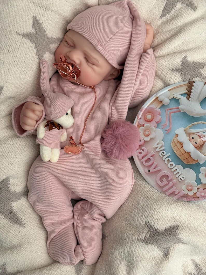 Pu&ograve; includere: Una bambola realistica che indossa una tutina rosa a coste a maniche lunghe con piedini e un berretto abbinato. La bambola ha un ciuccio in oro rosa, un piccolo peluche e un cartello rotondo con la scritta Welcome Baby Girl e una cicogna.