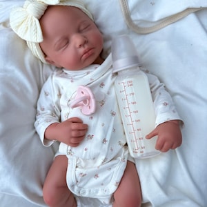 May include: A lifelike baby doll dressed in a cream onesie with a floral print, a matching bow headband, and white lace socks. A pink dummy and a clear baby bottle with milk are also present.