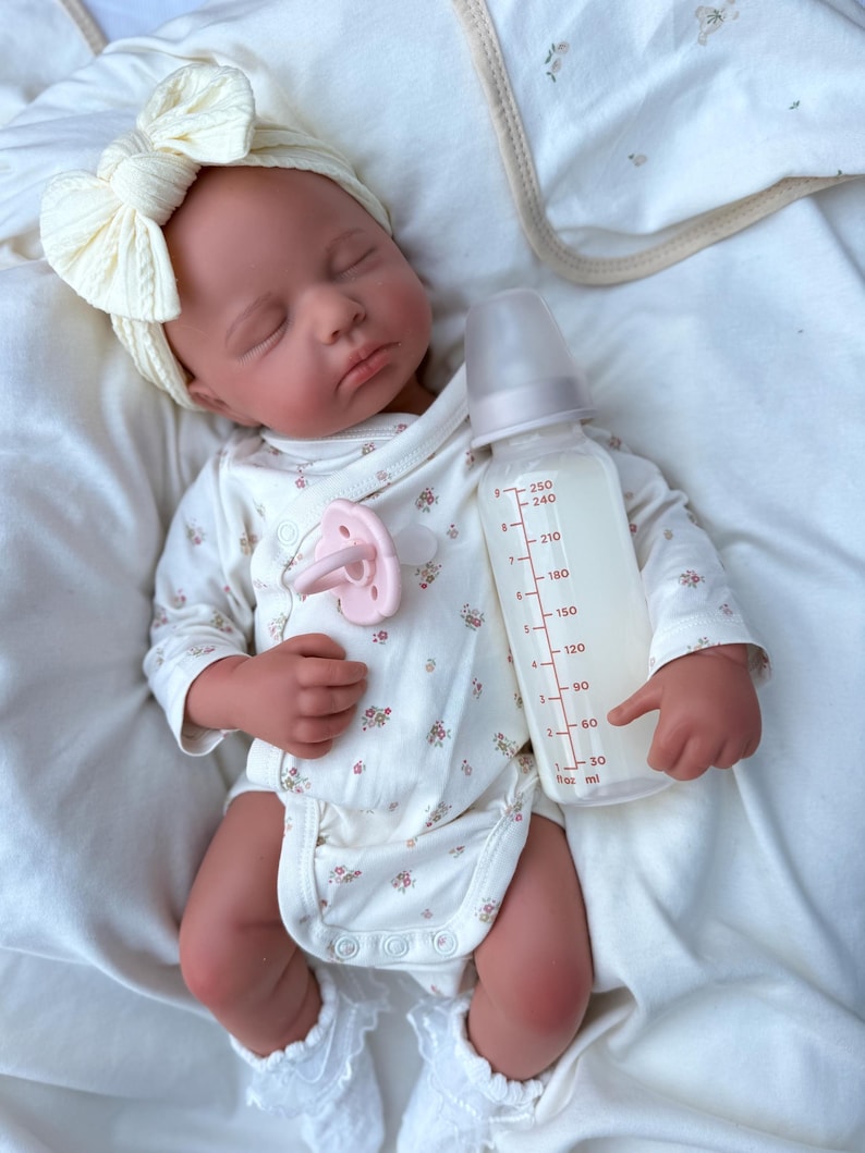 May include: A lifelike baby doll dressed in a cream onesie with a floral print, a matching bow headband, and white lace socks. A pink dummy and a clear baby bottle with milk are also present.