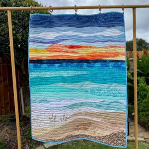 May include: A rectangular fabric quilt featuring a landscape scene with a sandy beach, turquoise ocean waves, and a vibrant sunset sky. The design includes horizontal wavy stitching patterns, patches of beach grass, and a pebble-textured border.