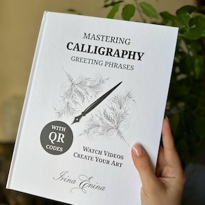 May include: A white softcover book titled Mastering Calligraphy Greeting Phrases by Irina Enina. The cover features a black ink illustration of a calligraphy pen and feathers, with text indicating the inclusion of QR codes for instructional videos.