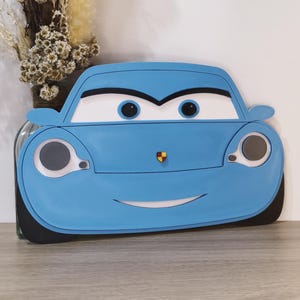 Può includere: Un oggetto decorativo a forma di auto azzurro con grandi occhi bianchi e pupille nere, una bocca bianca curva e un piccolo emblema sul cofano. Il design include fari circolari con centri grigi e specchietti laterali neri, simile a un personaggio.