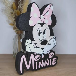 May include: A black Minnie Mouse figure with a pink bow and white polka dots. The figure has a white face and hands, with the name "Minnie" in pink letters at the base. The figure is on a wooden surface.